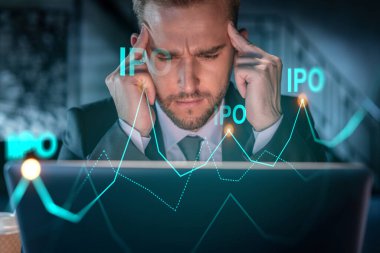 İş yerindeki yakışıklı iş adamı halka arz stratejisini optimize etmek için dizüstü bilgisayarla çalışıyor. Ofis arkaplanının finans şeması hologramı