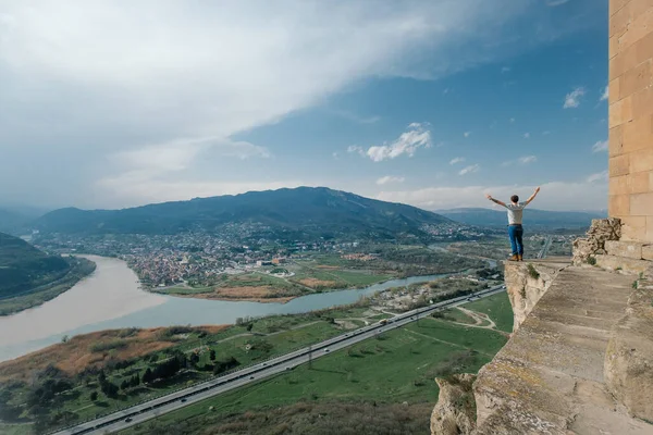 Bir adam yüksek bir dağın kenarında duruyor, kollarını farklı yönlere açıyor.