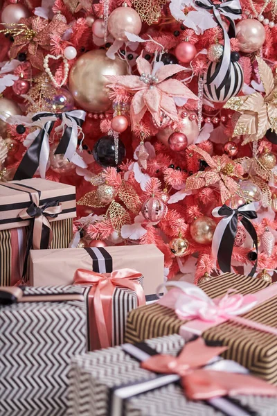 many boxes with gifts in black and white and pink paper with ribbons close-up lie under a very beautifully decorated pink Christmas tree with a lot of different Christmas toys.