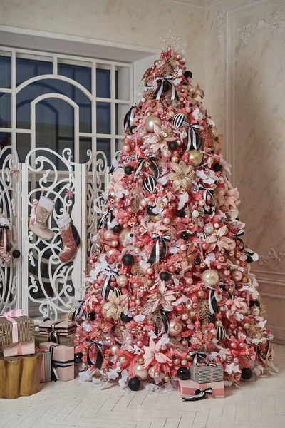 Tavana kadar birçok güzel oyuncakla ve toplarla süslenmiş uzun, güzel pembe bir Noel ağacı pencerenin önünde bir perdeyle duruyor. Ağacın altında hediyeler olan bir sürü kutu var.                               