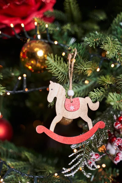 Christmas tree toy in the form of a wooden swinging horse with a red saddle hangs on the Christmas tree next to golden balls and garland lights