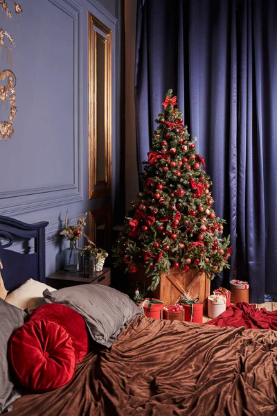 a beautiful Christmas tree decorated with red balls stands in the corner of the bedroom. blue curtains and walls are adorned with gold painting and frame. part of the bed is visible