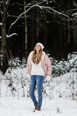 Beyaz eldivenli güzel bir kız, bir şapka ve açık renk giysiler içinde karlı bir ormanda gözleri kapalı ve yüzü gökyüzüne dikilmiş duruyor.