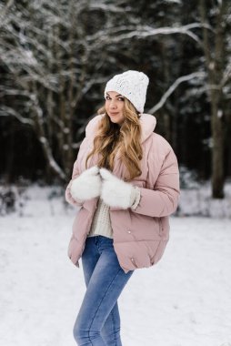 Kış kıyafetleri içinde uzun kıvırcık sarı saçlı ve karlı bir ormanda gülümseyen beyaz tüylü eldivenli güzel bir kadın.