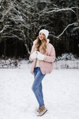 Kış kıyafetleri içinde uzun kıvırcık sarı saçlı ve karlı bir ormanda gülümseyen beyaz tüylü eldivenli güzel bir kadın.