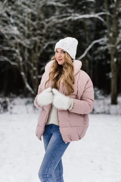 Kış kıyafetleri içinde uzun kıvırcık sarı saçlı ve karlı bir ormanda gülümseyen beyaz tüylü eldivenli güzel bir kadın.