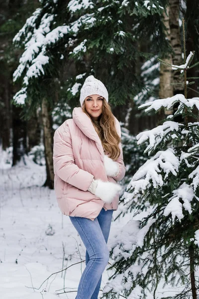 Beyaz şapkalı ve tüylü eldivenli güzel bir kız. Karlı bir ormanda ladin yanında pembe bir ceket.