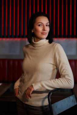  Güzel esmer kız dirseği yüksek bir sandalyede, çizgili bir duvarın önünde duruyor.                              