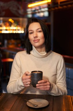 Omuz hizasında siyah saçlı güzel bir kız elinde bir kupayla bir kafede oturuyor.                               
