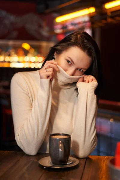 Zarif hatları olan, omuz hizasında koyu renk saçları olan, açık renk bir kazağı olan, boğazı olan, yüzünü bir kafede tasmasının arkasına saklayan güzel bir kız.                          