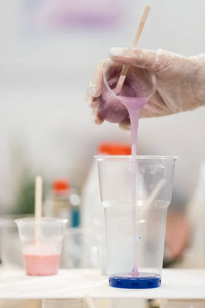 küçük bir bardaktan leylak reçinesi ve ahşap bir çubuktan şeffaf bir eldivene uzanan bir el ince bir reçine akışını mavi boyalı büyük şeffaf bir bardağa döküyor.