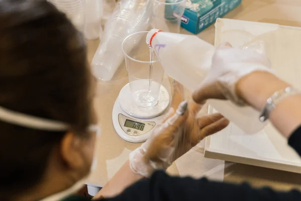 Maskeli ve eldivenli bir kız plastik şişeden şeffaf bir plastik bardağa temiz bir sıvı döküyor. Yukarıdan görünüm