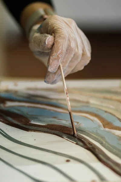 a gloved hand leads with a thin stick, creating waves and overflows in an epoxy painting, fluid art workshop