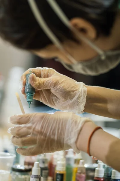 Maskeli ve şeffaf eldivenli bir kız. Renkli bir şişenin boyasıyla yakın plan bir şapka.