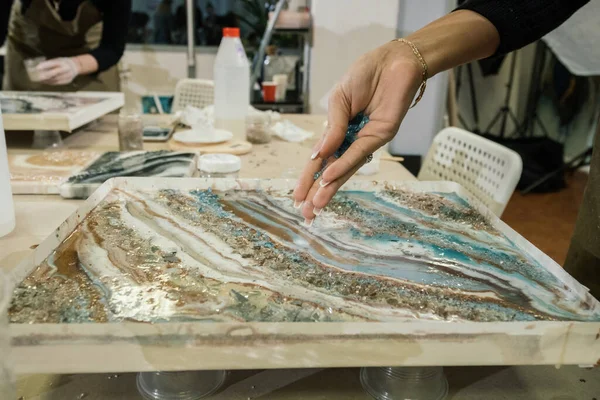 Sıvı sanat atölyesinde, uzun manikürlü bir kız, epoksi bir resmin üzerine sert mavi kırıntılar döker.