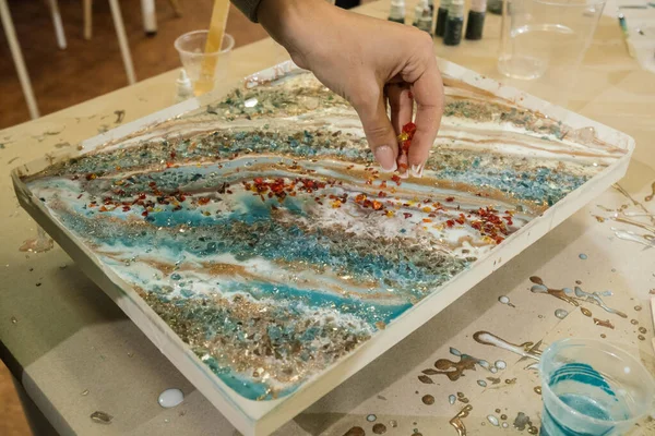 Sıvı sanat atölyesinde, uzun manikürlü bir kız, epoksi bir resmin üzerine sert kehribar kırıntıları döker.