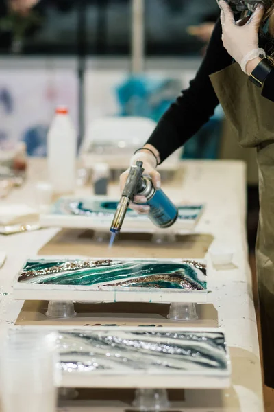 Eldivenli ve önlüklü bir kız bir elinde yanan bir ocağı tutarken, diğer elinde maske tutarak epoksi reçinesi resmi yaratıyor.