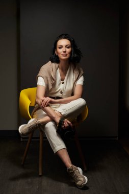Açık renk elbiseli güzel bir kadın koyu renkli bir koltukta oturuyor. Bir bacağı diğerinin üzerinde.                               
