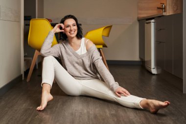 Açık renk elbiseli güzel, genç esmer kadın yere oturur, bir bacağını uzatır, dirseğini diğerinin dizine yaslar ve neşeyle gülümser.                             