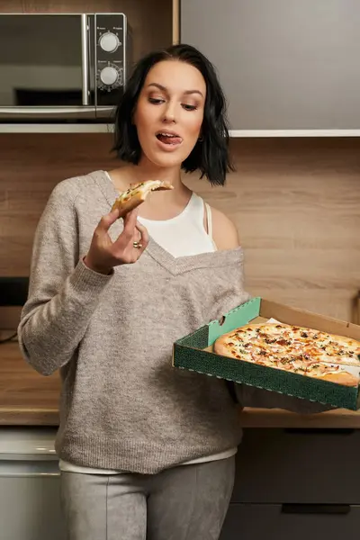 Koyu renk saçlı ve açık renk elbiseli genç bir kadın iştah açıcı bir şekilde dilini uzatmış ve elinde lezzetli bir dilim pizza tutuyor. Öte yandan bir kutu pizzayı elinde tutuyordu.                               