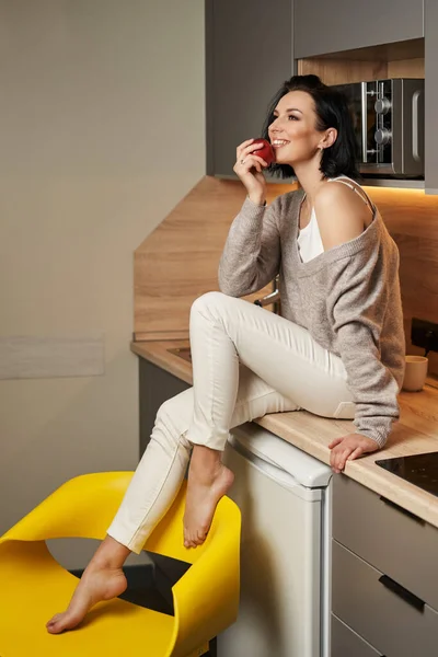 Çok güzel bir genç kadın mutfak tezgahında oturuyor, bacaklarını zarif bir şekilde sarı bir sandalyeye koyuyor ve yüzüne yakın güzel bir kırmızı elma tutuyor.                               