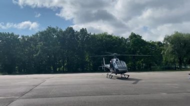 Kalkışa hazırlanan dönen bıçaklarla dolu büyük bir beton kalkış pistinde küçük özel bir helikopter. Ağaçlar arka planda yetişir. Gökyüzünde büyük bulutlar yüzüyor.