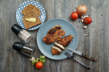 Bıçak ve çatalla tabakta iki kızarmış tavuk göğsü. Yanında tereyağlı iki dilim tost var. Proteinler karbonhidratları yağlar.