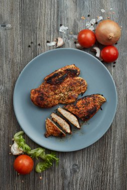 Gri bir tabakta, ahşap bir masada kızarmış tavuk göğsü. Yanında taze sebze, domates, soğan, sarımsak ve baharat var.