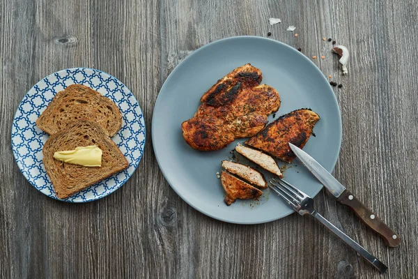 Bıçak ve çatalla tabakta iki kızarmış tavuk göğsü. Yanında tereyağlı iki dilim tost var. Proteinler karbonhidratları yağlar.