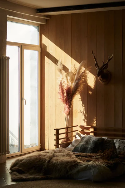 bedroom in Scandinavian style. bed on the floor. wooden interior. bouquet of tall pampas grass. large ceiling-to-floor window with sunlight
