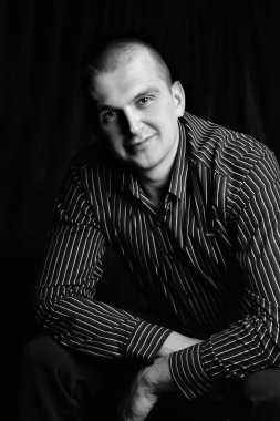 Stüdyoda elleri çapraz oturan, kameraya bakıp gülümseyen bir adamın siyah beyaz portresi.