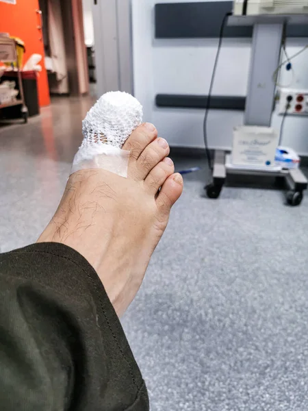 Vertical shot of a bare foot with the first toe bandaged to protect it, with gauze and a mesh in the form of a net, in a room and corridors of a hospital out of focus in the background.