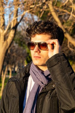 Güneş batımının altın saatinde güneş gözlüğü takan, palto ve eşarp takan kıvırcık saçlı beyaz bir çocuğun dikey çekimi.