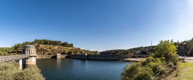 Tepeler ve bitki örtüsüyle çevrili bir barajın geniş panoramik manzarası, berrak mavi gökyüzünü yansıtan durgun su. Güçlü yatay kompozisyon ve cömert kopya alanına sahip doğal manzarayı aç.