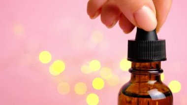 Pembe arka planda cilt bakımı kozmetik yağ şişesi. Kadın eli bir pipeti tutuyor ve yağ damlatıyor. Alternatif ilaç, C vitamini, çiçek suyu ve kozmetik serum. Koyu kahverengi cam.