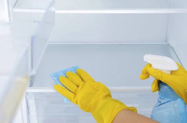 Female hands in a yellow rubber protective glove and a blue sponge washes, cleans refrigerator shelves. Cleaning service, housewife, routine housework. Spray for windows and glass surfaces cleaner Stock Image