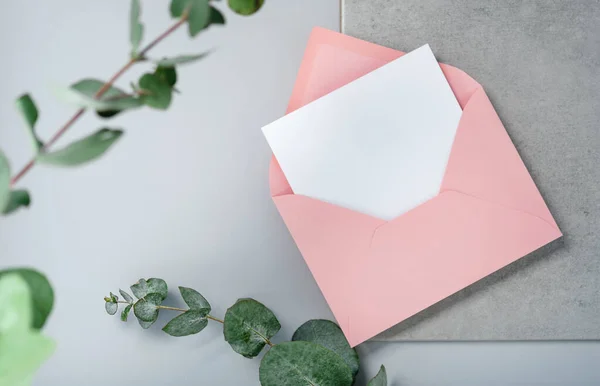 Real photo. Pink envelope square invitation card mockup with a eucalyptus branch. Top view with copy space, light gray background. Template for branding and advertising