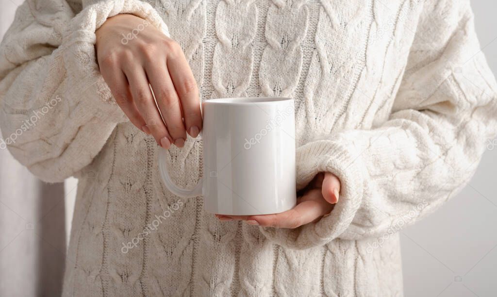 Female hands hold mock up white empty mug, cup for your design and logo close-up. Woman in beige warm knitted sweater autumn winter. Blank template for promotional text message or promotional content