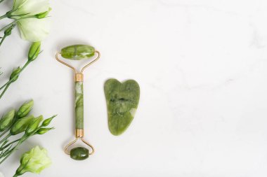 Yüz masajı için Jade Roller. Yeşil gua sha yüz masajı araçları mermer beyaz arka planda fotokopi alanı düz yatıyordu. Yaş, kaldırma ve tonlama tedavisi, akupres. Eustoma çiçekleri.