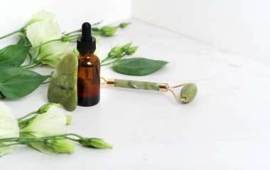 Yüz masajı için Jade Roller. Yeşil gua sha mermer beyaz arka planda düz yatıyordu. Yaş, kaldırma ve tonlama tedavisi, akupres. Eustoma çiçekleri. Pipetli serum kahverengi şişe..