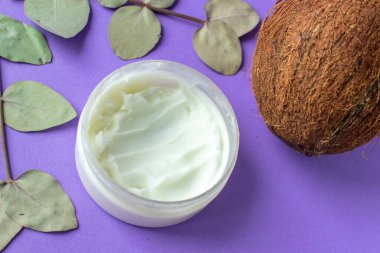 Hindistan cevizi yağlı kavanozda hindistan cevizi ve mor arka planda yapraklar.