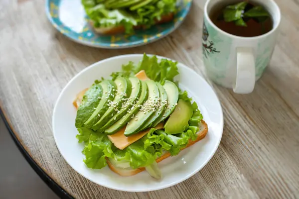 Avokado toast på vetebröd med ost, sallad och sesamfrön på en vit tallrik på ett träbord. Sund frukost. — Stockfoto