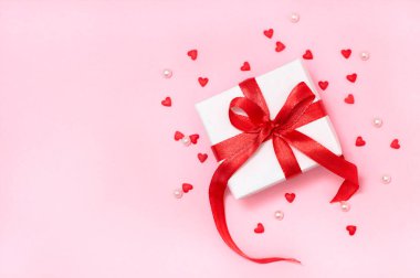 Kırmızı kurdeleli bir hediye kutusu, küçük şeker kalpler ve son moda pembe arka planda beyaz boncuklar. Düz yatış, üst görünüm Sevgililer Günü afişi veya tebrik kartı