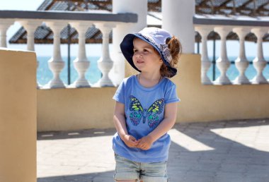 Küçük gülümseyen kız, Yunan tarzında deniz manzaralı taş bir duvarın yanında duruyor.