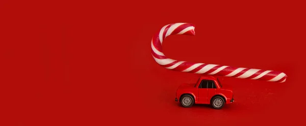 Świąteczna makieta. Koncepcja samochodu do dostarczania prezentów świątecznych. Czerwony samochód niesie cukierkową laskę na czerwonym tle. Widok z przodu kreatywna abstrakcyjna kompozycja banerowa z miejscem na tekst. — Zdjęcie stockowe
