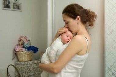 Anne yeni doğmuş bebeğini evde bir çamaşır sepetinin arka planında nazikçe öpüyor. Anne çocuğu nazikçe kollarına aldı. Ev işleri ve yalnız bir annenin endişeleri. Kopyalama alanı