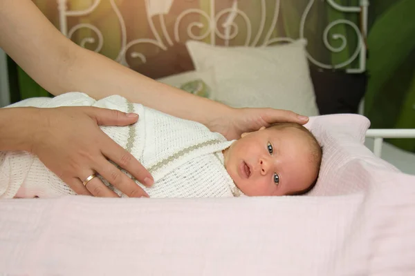 Женские руки пеленают ребенка в белом покрывале. Мама гладит ребенка, который лежит дома на кровати и смотрит в рамку. Концепция семьи, детства, домашнего рождения, новой жизни. Copyspace — стоковое фото