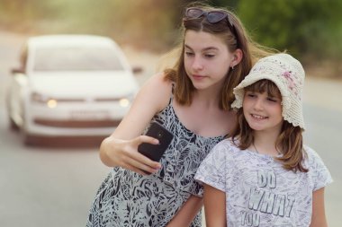Çocuklar yolda dururken, tehlikeyi fark etmeden cep telefonuyla selfie çekiyorlar. Hareket halindeki beyaz bir arabanın arka planında iki kızın fotoğrafı çekiliyor..