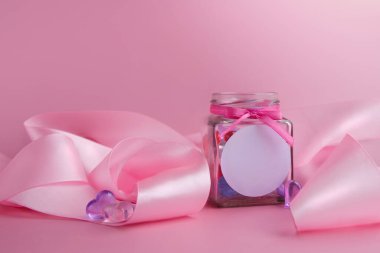 Logo için bir yeri olan bir maket. İçinde kalpler olan cam kavanoz ve pembe arka planda pembe ipek kurdele. Aşk konsepti ve Sevgililer Günü için. Banner, şablon. Yatay format. Metin için yer.
