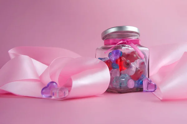 ピンクの背景にキャンディとピンクのシルクリボンでハート型のガラス瓶。バレンタインデーのコンセプト。サイドビュー。水平形式。コピースペース — ストック写真